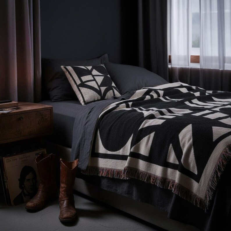 Black and cream geometric woven blanket with multicolor fringed edges styled on a dark-toned bed with a matching pillow. The dimly lit room features wooden accents, leather boots, and a candle, creating a moody, modern interior atmosphere.