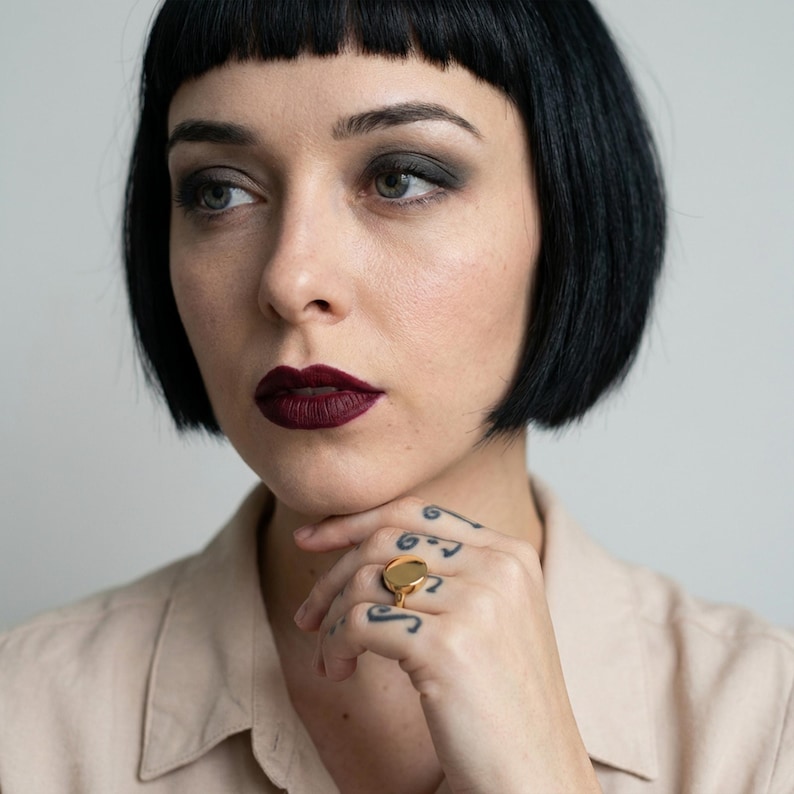 close portrait with soft, natural makeup and deep lip color, gaze turned slightly off-camera, hand resting under the chin with a gold signet ring, fine finger tattoos adding detail, clean background with gentle, even light