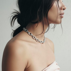 Close-up of a woman wearing a silver sculptural chain necklace with oval open links. The necklace rests along the collarbone, styled with a soft strapless top. Minimal, modern jewelry with an organic industrial design captured in natural light.