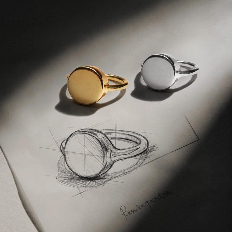 gold and silver round signet rings placed above a hand-drawn design sketch, soft directional light creating contrast and depth, clean geometric form echoed in the drawing below, a quiet Rannka Poetcore collection study of form and material
