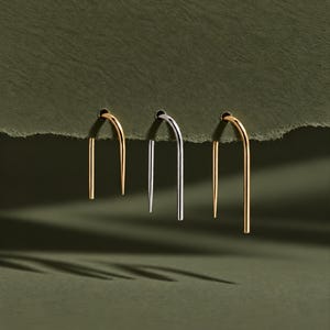 May include: Three U-shaped earrings hanging from a textured edge. The left earring is gold, the center earring is silver, and the right earring is gold. Each earring features a smooth, curved top and two straight, parallel vertical bars.