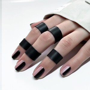 以下が含まれることがあります： 黒い革の指輪のクローズアップ。黒とピンクのマニキュアが施された指に装着されています。リングは幅広の革バンドです。爪はピンクの背景に斜めの黒いストライプでペイントされています。
