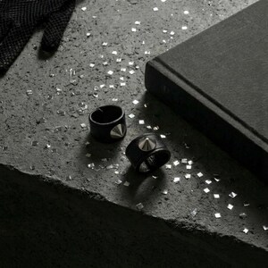 Black vegan leather stud rings with silver cone hardware styled on a raw concrete surface. Two minimalist leather band rings placed beside a dark fabric-covered jewelry box, scattered with small metallic confetti pieces. Moody, industrial flat lay with matte textures and soft shadows. Edgy unisex ring design, modern alternative jewelry styling, Rannka aesthetic.