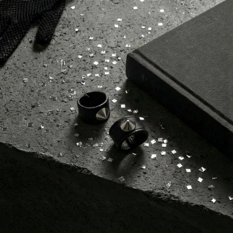 Black vegan leather stud rings with silver cone hardware styled on a raw concrete surface. Two minimalist leather band rings placed beside a dark fabric-covered jewelry box, scattered with small metallic confetti pieces. Moody, industrial flat lay with matte textures and soft shadows. Edgy unisex ring design, modern alternative jewelry styling, Rannka aesthetic.