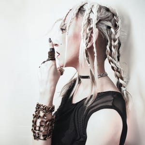 young woman posing on a side with her finger to her lips, wearing black mesh elegant dress and statement jewelry, in particular a large gold finish statement ring. her hair is in large chunky braids.