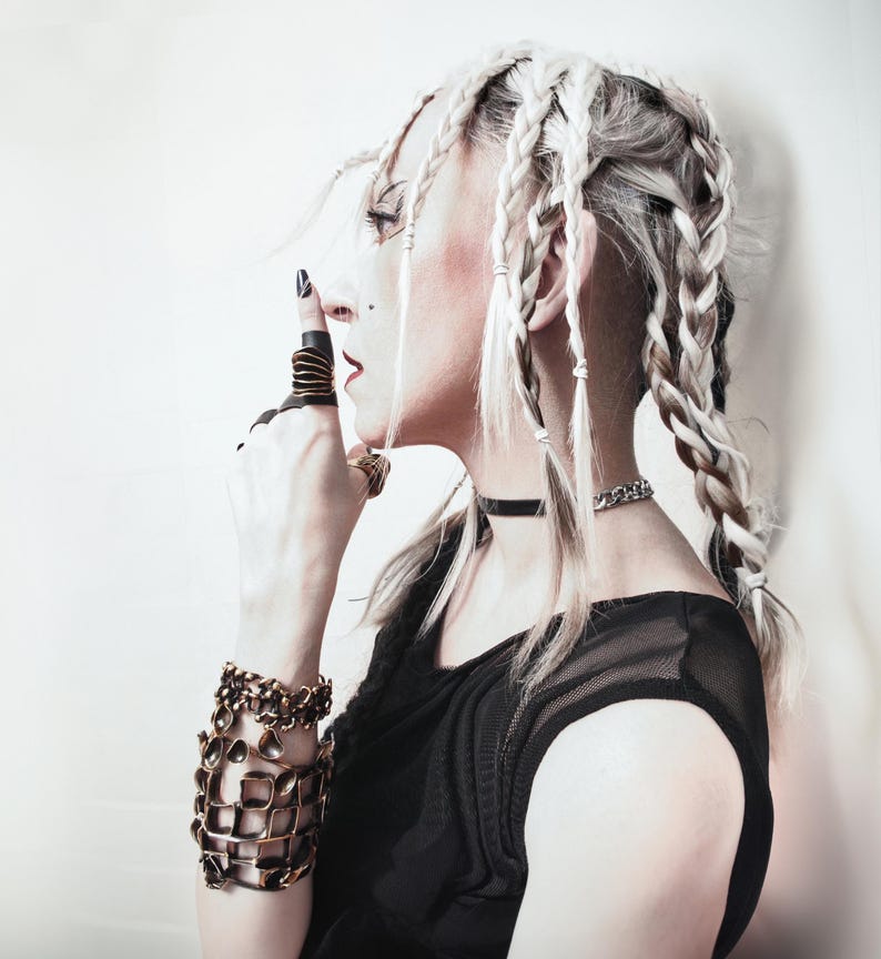 young woman posing on a side with her finger to her lips, wearing black mesh elegant dress and statement jewelry, in particular a large gold finish statement ring. her hair is in large chunky braids.