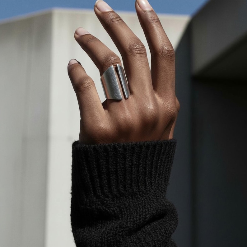 Close-up of a hand wearing a bold silver geometric ring with a wide open-ended design, paired with a black knit sleeve against a soft architectural background.