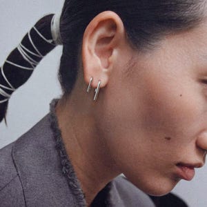 Sterling silver linear drop earrings in small and medium lengths worn as a double piercing set, sleek vertical bar earrings with clean lines, minimalist ear stack styling, modern geometric jewelry look, subtle everyday silver earrings, close-up side profile with tailored jacket, Rannka aesthetic