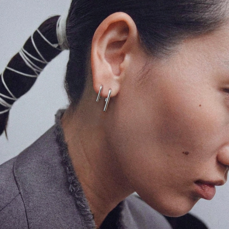 Sterling silver linear drop earrings in small and medium lengths worn as a double piercing set, sleek vertical bar earrings with clean lines, minimalist ear stack styling, modern geometric jewelry look, subtle everyday silver earrings, close-up side profile with tailored jacket, Rannka aesthetic