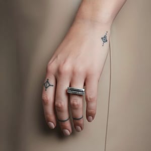 Hand wearing a geometric silver bar ring with a raw hammered finish. Minimalist design with clean lines, styled with small tattoos and neutral tones. Adjustable unisex ring ideal for stacking or everyday wear. Rannka modern jewelry.