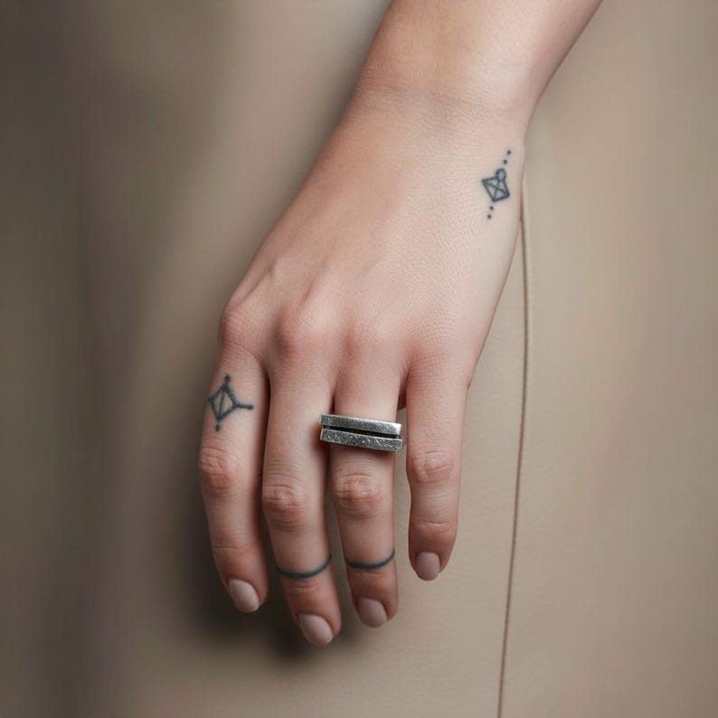 Hand wearing a geometric silver bar ring with a raw hammered finish. Minimalist design with clean lines, styled with small tattoos and neutral tones. Adjustable unisex ring ideal for stacking or everyday wear. Rannka modern jewelry.
