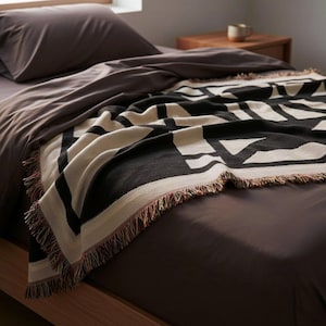 Black and cream geometric woven blanket with multicolor fringed edges draped over a neatly made bed with brown sheets. The textured pattern adds depth and structure to a minimal modern bedroom with soft morning light.