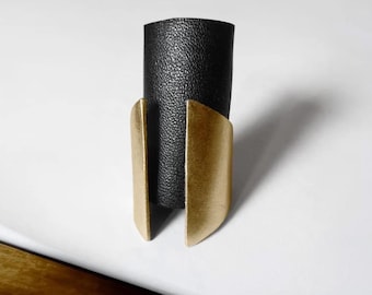 May include: A black leather cuff bracelet with a gold-colored metal accent. The bracelet has a textured leather band and a curved, open metal design on the front. The cuff is set against a white background.