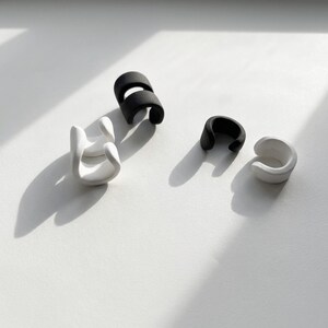 Black and white ear cuff pieces arranged on a light surface, no piercing ear jewelry set showing individual matte ear wraps, minimalist sculptural design with soft shadows, modern stacked ear accessories photographed flat lay, contemporary Rannka jewelry styling.