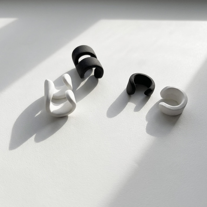 Black and white ear cuff pieces arranged on a light surface, no piercing ear jewelry set showing individual matte ear wraps, minimalist sculptural design with soft shadows, modern stacked ear accessories photographed flat lay, contemporary Rannka jewelry styling.