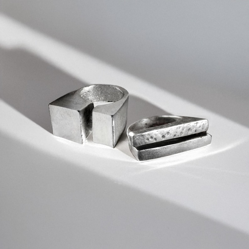 Two brutalist silver rings on a white surface with sharp light and shadow. One features a split-square architectural band, the other stacked geometric layers with raw hammered texture. Contemporary sculptural jewelry by Rannka, modern industrial style statement rings, bold and minimal design.
