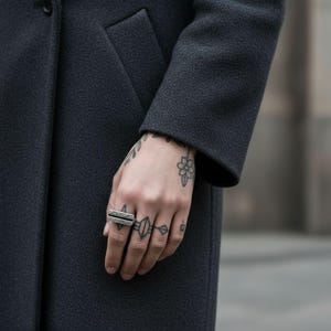 Hand wearing a geometric hammered silver bar ring with a raw, structured design. Styled with black tattoos and a dark wool coat. Modern industrial piece for men or women, combining minimalist and streetwear aesthetics. Rannka jewelry.