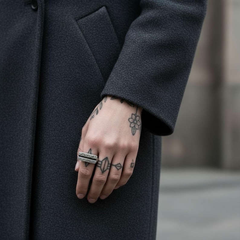Hand wearing a geometric hammered silver bar ring with a raw, structured design. Styled with black tattoos and a dark wool coat. Modern industrial piece for men or women, combining minimalist and streetwear aesthetics. Rannka jewelry.