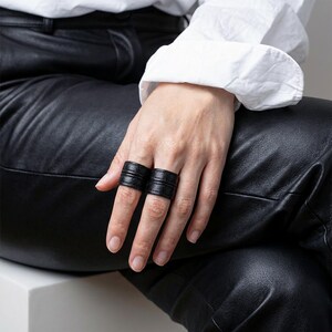 Close-up of a hand wearing two black vegan leather wrap rings, styled across adjacent fingers while resting on black leather pants. Minimal leather ring cuffs with layered band detail and matte texture, shown worn to highlight scale, fit, and everyday styling. Clean editorial composition with soft natural light and a white cuffed sleeve for contrast. Contemporary cruelty-free jewelry by Rannka, designed for modern ring stacking, understated statement wear, and thoughtful gifting.