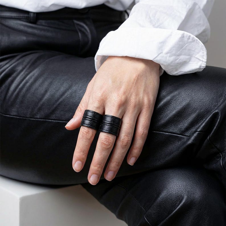 Close-up of a hand wearing two black vegan leather wrap rings, styled across adjacent fingers while resting on black leather pants. Minimal leather ring cuffs with layered band detail and matte texture, shown worn to highlight scale, fit, and everyday styling. Clean editorial composition with soft natural light and a white cuffed sleeve for contrast. Contemporary cruelty-free jewelry by Rannka, designed for modern ring stacking, understated statement wear, and thoughtful gifting.