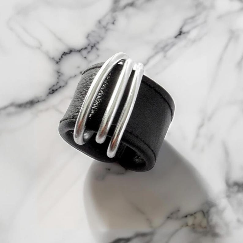 Avant-Garde Black Leather Ring, Black vegan leather ring with three silver metal bands wrapped across the front, photographed on a marble surface, modern mixed-material accessory design.