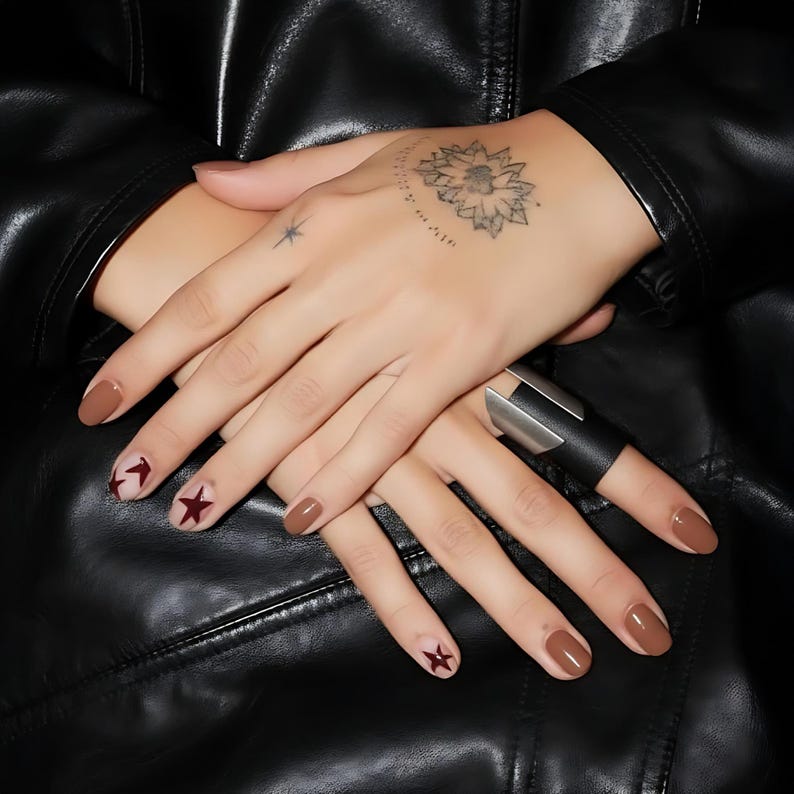 Close-up of hands with brown and burgundy star nail art, wearing a geometric black vegan leather and steel ring on the index finger. The hands rest on a black leather jacket, showing minimalist tattoos and neutral modern styling. Fashion editorial aesthetic, moody lighting, and edgy accessory focus.