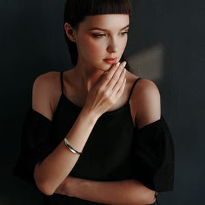 May include: A silver-colored, curved bracelet is worn on the left wrist. The bracelet has a smooth, polished surface. The person in the image is wearing a black spaghetti strap top and a black off-the-shoulder top.