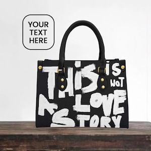 Girlfriend Initials Gift Idea Evening Bag, Black handbag with bold white painted text reading “THIS IS NOT A LOVE STORY,” featuring black handles with gold hardware, displayed on a rustic wooden table against a minimal white wall background.