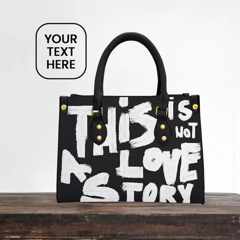Girlfriend Initials Gift Idea Evening Bag, Black handbag with bold white painted text reading “THIS IS NOT A LOVE STORY,” featuring black handles with gold hardware, displayed on a rustic wooden table against a minimal white wall background.