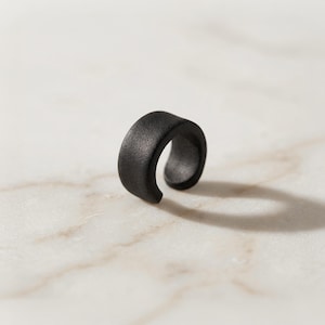 Matte black ear cuff displayed alone on light stone surface, no piercing ear jewelry with curved open design, minimalist ear wrap for everyday wear, modern sculptural ear accessory shown as single piece, contemporary handmade ear jewelry by Rannka.