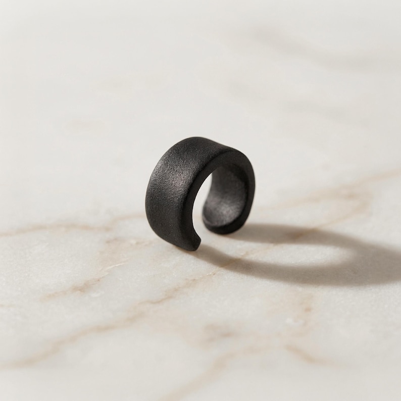Matte black ear cuff displayed alone on light stone surface, no piercing ear jewelry with curved open design, minimalist ear wrap for everyday wear, modern sculptural ear accessory shown as single piece, contemporary handmade ear jewelry by Rannka.