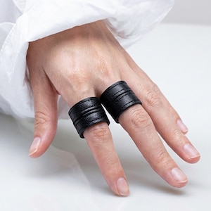 May include: Two black leather finger rings. Each ring is a wide band with horizontal textured lines. The rings are worn on the index and middle fingers. The background is a white shirt.
