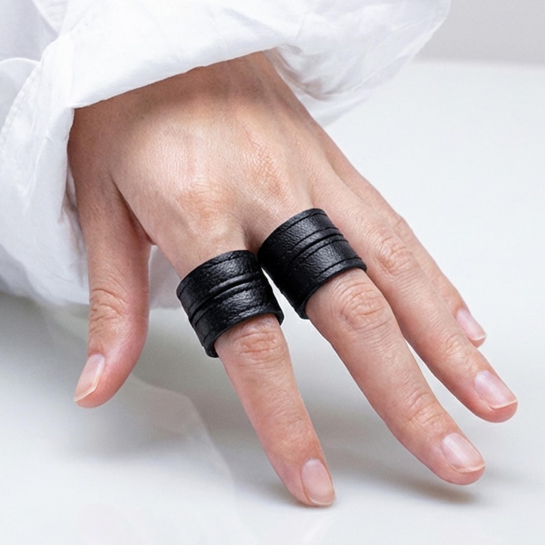 Two black vegan leather wrap rings worn stacked across adjacent fingers, photographed on a hand against a clean white background. Minimal leather ring cuffs with layered band detail and matte texture, shown worn to demonstrate scale, fit, and styling. Contemporary cruelty-free jewelry with an architectural, understated look, styled in a modern editorial way. Designed by Rannka for everyday wear, ring stacking, and giftable minimalist styling.