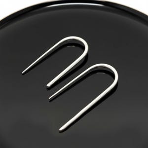 Sterling silver U shape earrings on black surface, minimalist open hoop earrings with tapered ends, modern sculptural ear wire design, sleek geometric silver jewelry, clean industrial style accessory, simple everyday earrings, Rannka style, high contrast product shot