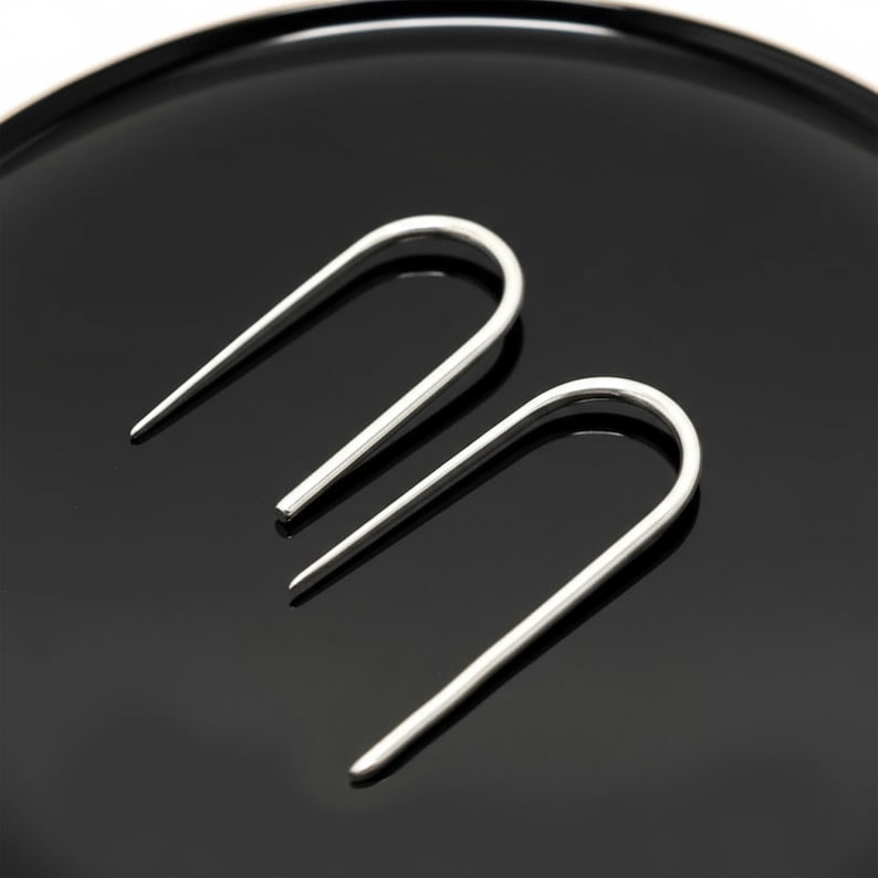 Sterling silver U shape earrings on black surface, minimalist open hoop earrings with tapered ends, modern sculptural ear wire design, sleek geometric silver jewelry, clean industrial style accessory, simple everyday earrings, Rannka style, high contrast product shot
