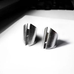 Pair of adjustable metal rings with sculptural split-front design and matte brushed finish. Open band construction with curved, folded metal panels creating an architectural, industrial look. Minimal contemporary unisex rings photographed on a clean white surface with strong light and shadow contrast. Raw texture and bold form emphasized, by Rannka.