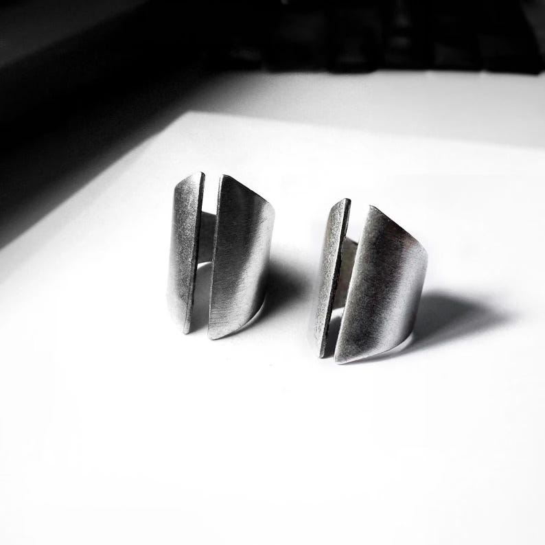 Pair of adjustable metal rings with sculptural split-front design and matte brushed finish. Open band construction with curved, folded metal panels creating an architectural, industrial look. Minimal contemporary unisex rings photographed on a clean white surface with strong light and shadow contrast. Raw texture and bold form emphasized, by Rannka.