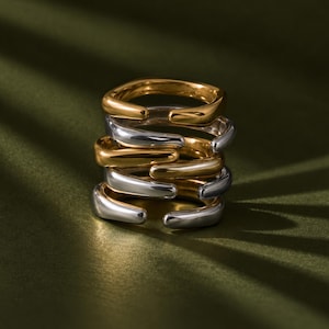 May include: A stack of five open-ended rings featuring alternating gold and silver-toned metal finishes. The rings have a smooth, rounded, and slightly irregular shape, creating a layered appearance with visible gaps at the front of each band.