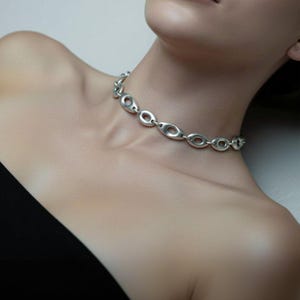 Close-up of a woman wearing a silver sculptural oval-link necklace styled as a choker against bare skin and a black strapless top. The smooth open links and reflective finish give it a modern, architectural look — minimal yet bold jewelry design.