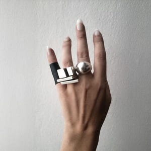 Hand wearing bold sculptural rings, including a brutalist silver geometric ring paired with a smooth dome ring and a wide black band. Contemporary architectural jewelry design by Rannka, edgy statement style with contrasting shapes and textures, modern industrial aesthetic for everyday or artistic wear.