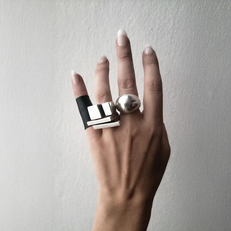 Hand wearing bold sculptural rings, including a brutalist silver geometric ring paired with a smooth dome ring and a wide black band. Contemporary architectural jewelry design by Rannka, edgy statement style with contrasting shapes and textures, modern industrial aesthetic for everyday or artistic wear.