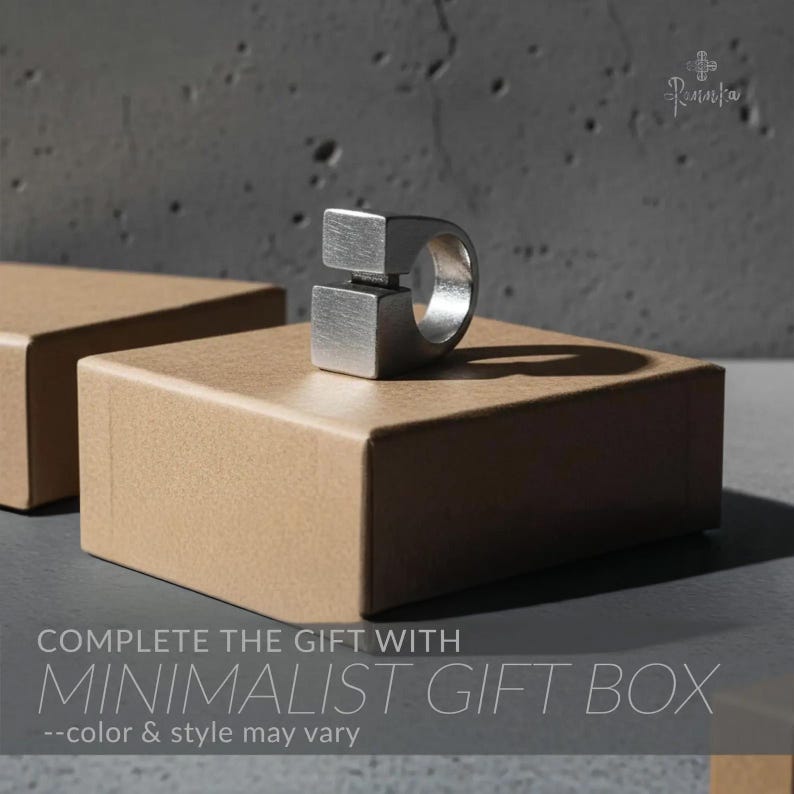 May include: A minimalist silver ring with a geometric design, featuring two square blocks connected by a band. The ring is displayed on a brown gift box. The text "COMPLETE THE GIFT WITH MINIMALIST GIFT BOX" is visible.