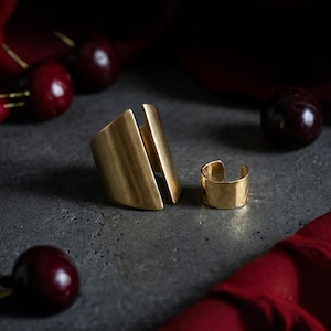 Sculptural gold ring by Rannka shown as the main focus, paired with a matching 14k gold filled ear cuff and bold gold wrap ring, arranged on a raw concrete surface with deep red accents, styled in a modern editorial setting highlighting Rannka’s minimalist handcrafted jewelry aesthetic.