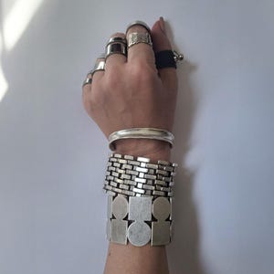 Hand wearing multiple silver rings and three chunky silver bracelets, displayed against a plain light gray background with natural lighting and subtle shadows, showcasing the bold, textured, and geometric designs of the jewelry.