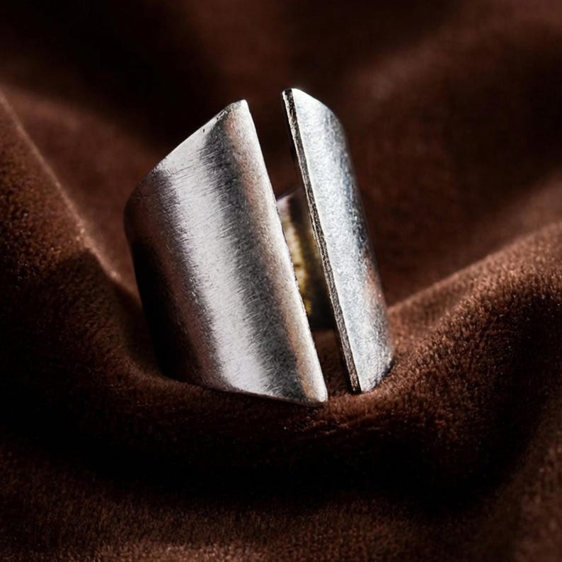 Silver split band minimalist ring on brown velvet background, Rannka style; open front design with brushed metal texture; close-up jewelry shot showing raw, organic finish and sculptural shape; moody soft lighting highlighting the ring’s edges and form; modern unisex metal ring aesthetic.