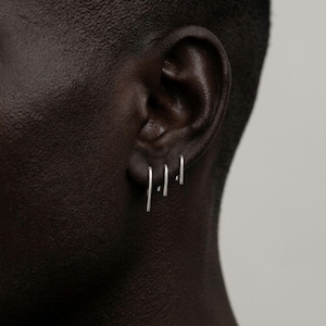 Minimalist sterling silver slip earrings on ear, small and medium bar drop earrings in clean stacked piercings, modern asymmetric ear styling, sleek industrial jewelry look, subtle everyday silver earrings, close-up ear detail, Rannka style, understated and sculptural mood
