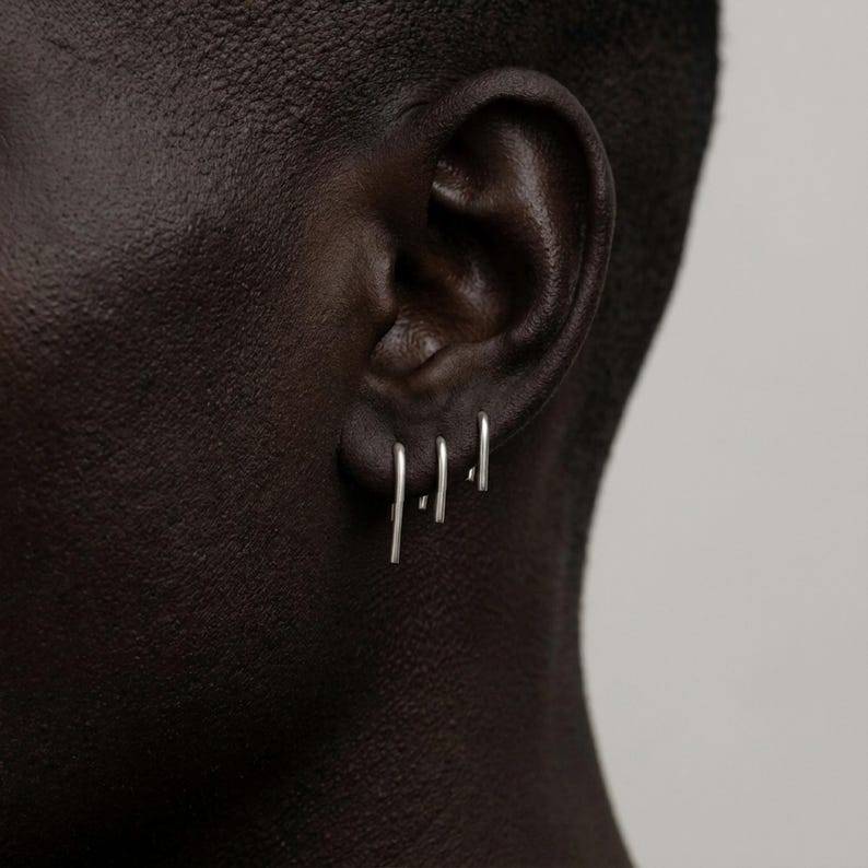 Minimalist sterling silver slip earrings on ear, small and medium bar drop earrings in clean stacked piercings, modern asymmetric ear styling, sleek industrial jewelry look, subtle everyday silver earrings, close-up ear detail, Rannka style, understated and sculptural mood