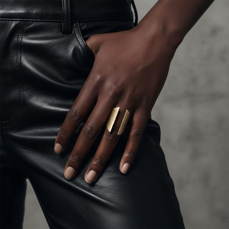 Gold open-band ring with sculptural split design worn on the hand, paired with black leather pants, minimalist modern styling against neutral background, contemporary jewelry piece by Rannka.