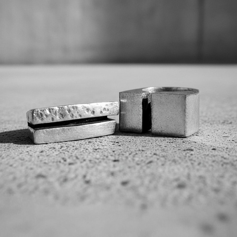 Close-up of two silver rings on a concrete surface — one with a hammered split-bar form, the other a solid open-square design. Matte finish highlights the raw metal texture. Industrial modern jewelry for both men and women. Rannka design.