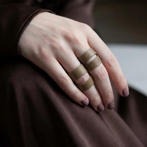 May include: Close-up of two brown leather rings on a hand. The rings have a unique design with cut-out sections. The hand has manicured nails painted in a matching brown color. The background is a blurred brown fabric.